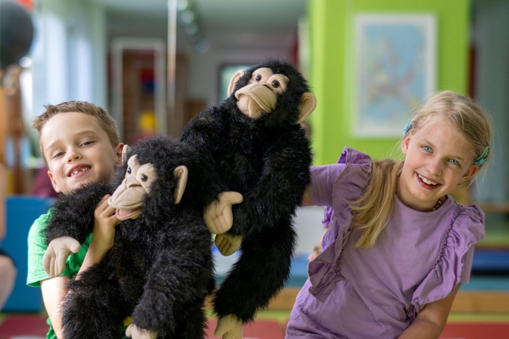 Ein lächelnder Junge und ein lächelndes Mädchen halten jeweils eine Schimpansen-Handpuppe hoch. Der Junge trägt ein grünes Hemd und das Mädchen ein lila Oberteil.