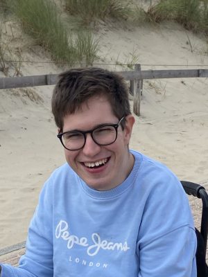 Ein junger Mann mit kurzen braunen Haaren und einer schwarzen Brille lächelt breit in die Kamera. Er trägt einen hellblauen Pullover.