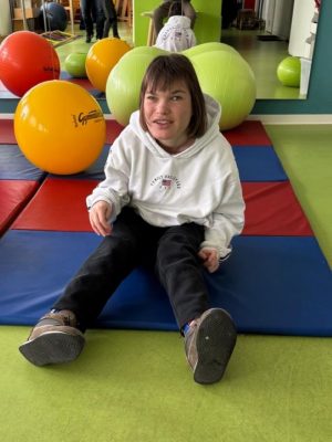 Eine junges Mädchen mit einem weißen Kapuzenpulli sitzt auf einer roten und blauen Matte in einem Raum mit bunten Gymnastikbällen.