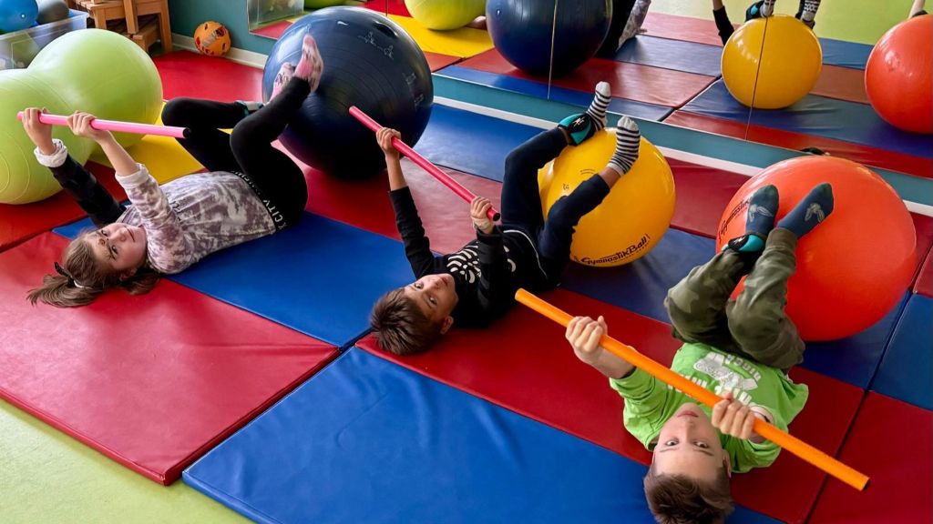 Drei Kinder liegen in unseren Räumlichkeiten auf dem Rücken auf roten und blauen Matten. Sie halten jeweils eine Stange mit beiden Händen und stützen ihre Beine auf großen, runden Bällen.