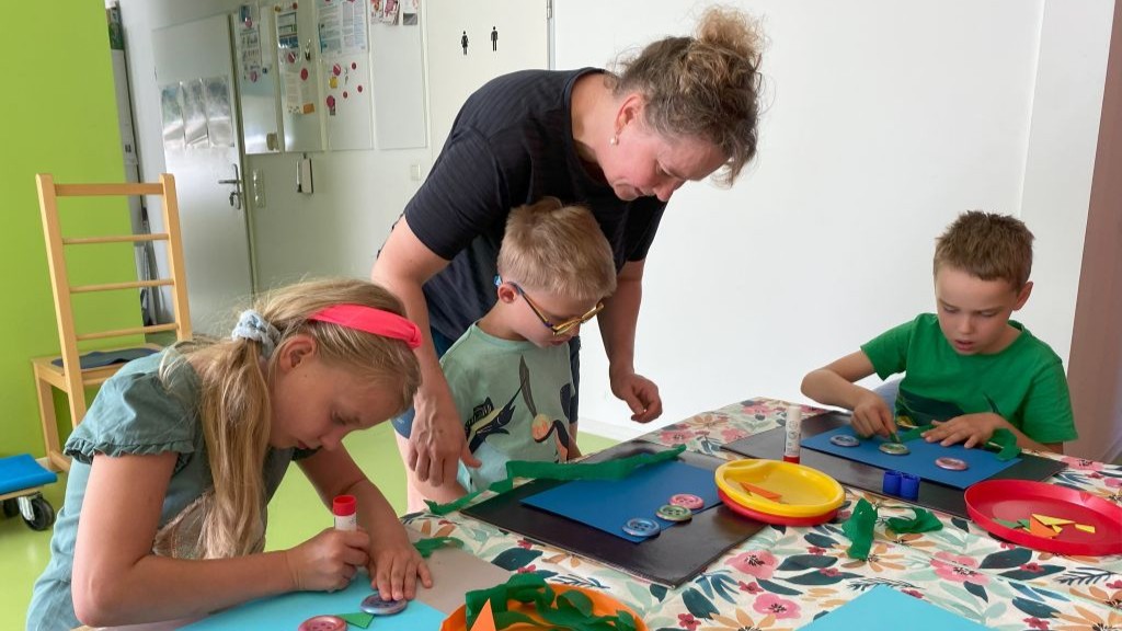 Unsere Konduktorin Eni lehnt sich über einen Tisch, um einem Jungen beim Basteln zu helfen, während ein Mädchen und ein anderer Junge an ihren eigenen Projekten arbeiten. Auf dem Tisch befinden sich verschiedene Bastelmaterialien.