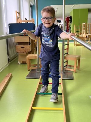 Ein lächelnder Junge mit Brille steht in einem Laufbarren und hält sich fest.