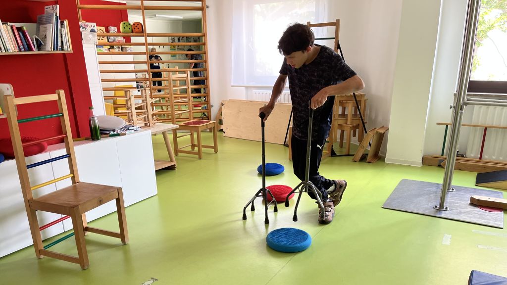 Ein junger Mann mit dunklen Haaren und einem dunklen T-Shirt stützt sich auf zwei Gehstöcke. Er steht auf einem hellgrünen Boden und bewegt sich vorsichtig um drei runde Balancekissen herum, die auf dem Boden verteilt sind.