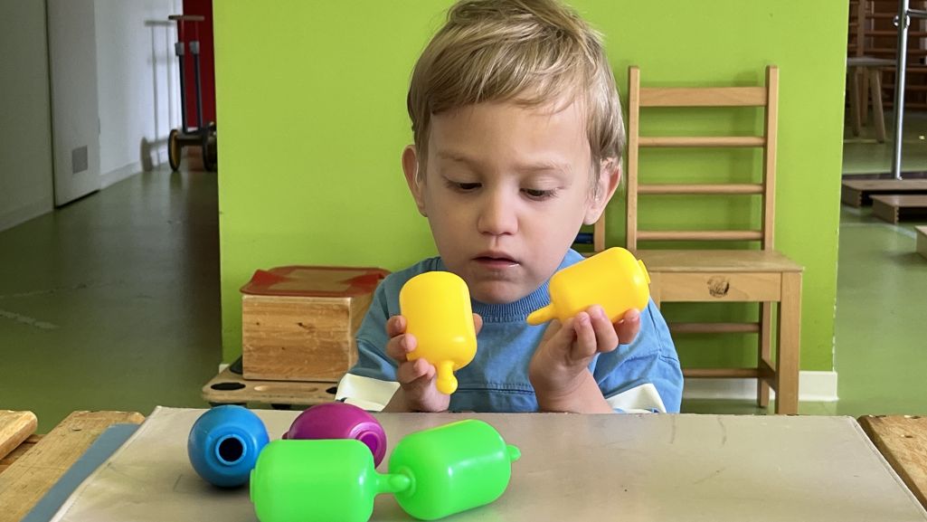 Ein Kind mit blonden Haaren in einem hellblauen T-Shirt, sitzt an einem Tisch und steckt konzentriert zwei gelbe, zylindrische Steckspielzeugteile ineinander.
