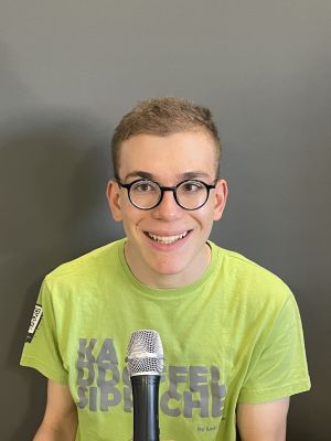 Portrait eines lächelnden, jungen Mannes mit kurzem Haar und runder Brille. Er sitzt vor einem Mikrofon und blickt direkt in die Kamera.