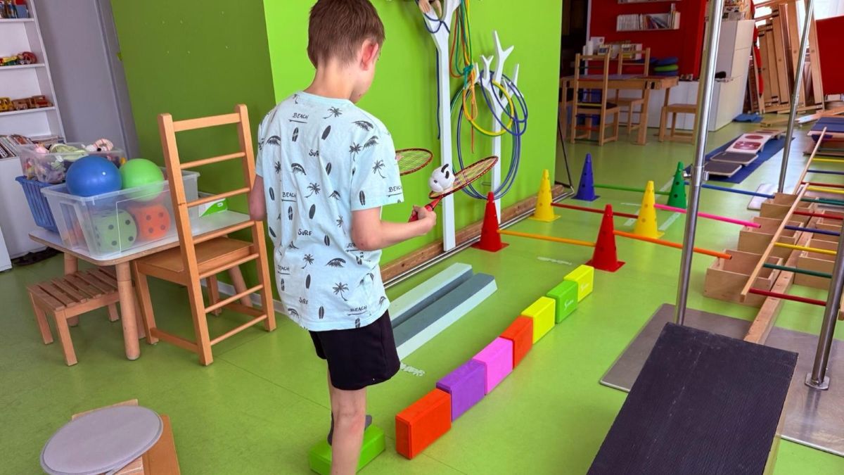 Kind in einem Therapieraum, das seine motorischen Fähigkeiten trainiert, indem es einen Ball mit zwei Badminton-Schlägern über einen Balancierparcours hält.